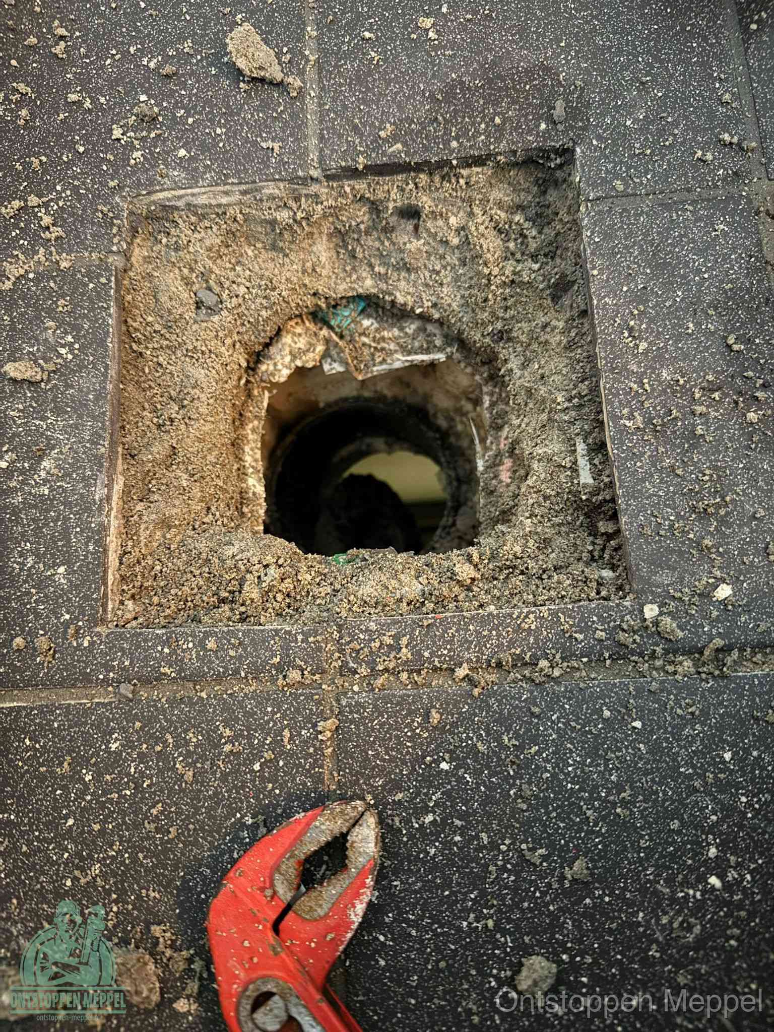 Een geopend rioolputje in een betontegelvloer met losgemaakt rooster en een zichtbare ronde afvoerbuis, het achterhalen waar de afvoerverstopping zit in Meppel