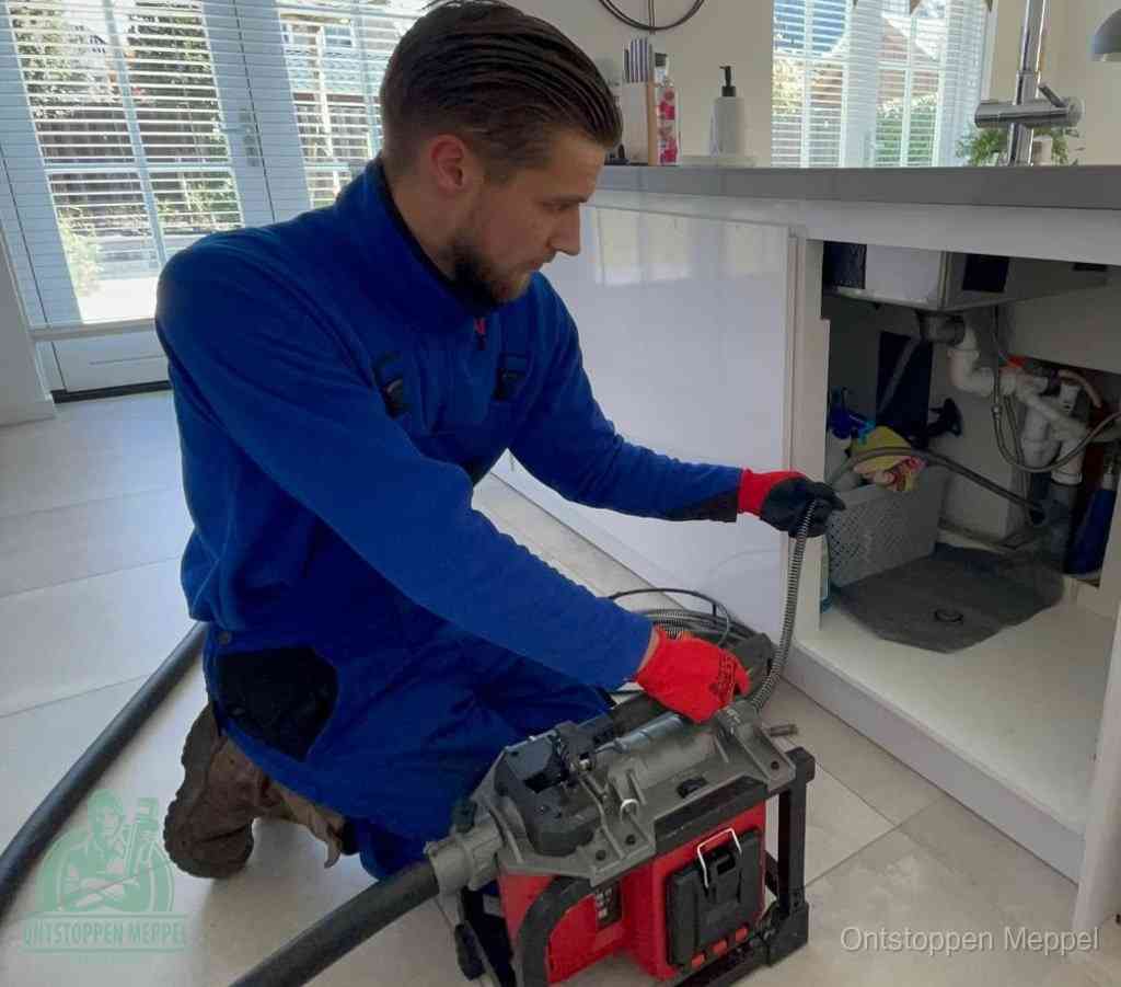 Een monteur met rode handschoenen knielt in een keuken en voert een elektrische ontstoppingsveer in tijdens het professioneel afvoer ontstoppen in Meppel